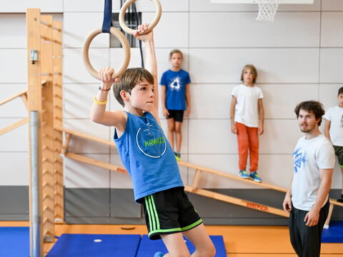 Ein Junge hängt an Turnringen in der Halle, ein Trainer und weitere Kinder schauen zu.
