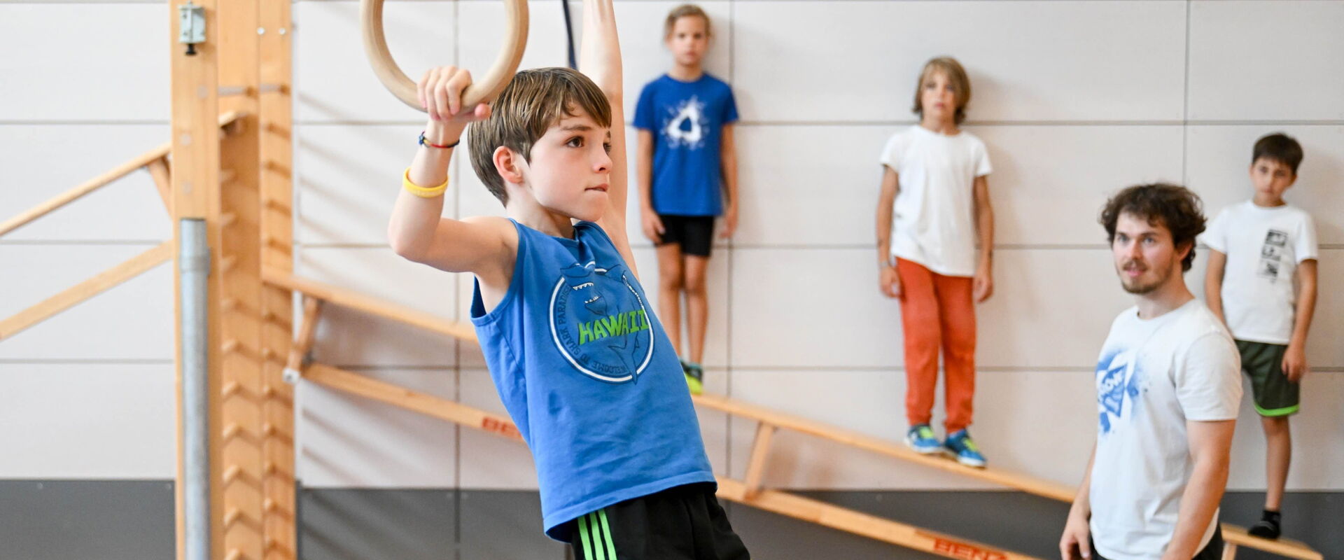 Ein Junge hängt an Turnringen in der Halle, ein Trainer und weitere Kinder schauen zu.