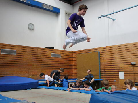 Ein Sprung hoch über einem Trampolin in der Halle, wartende Kinder sitzen am Rand.